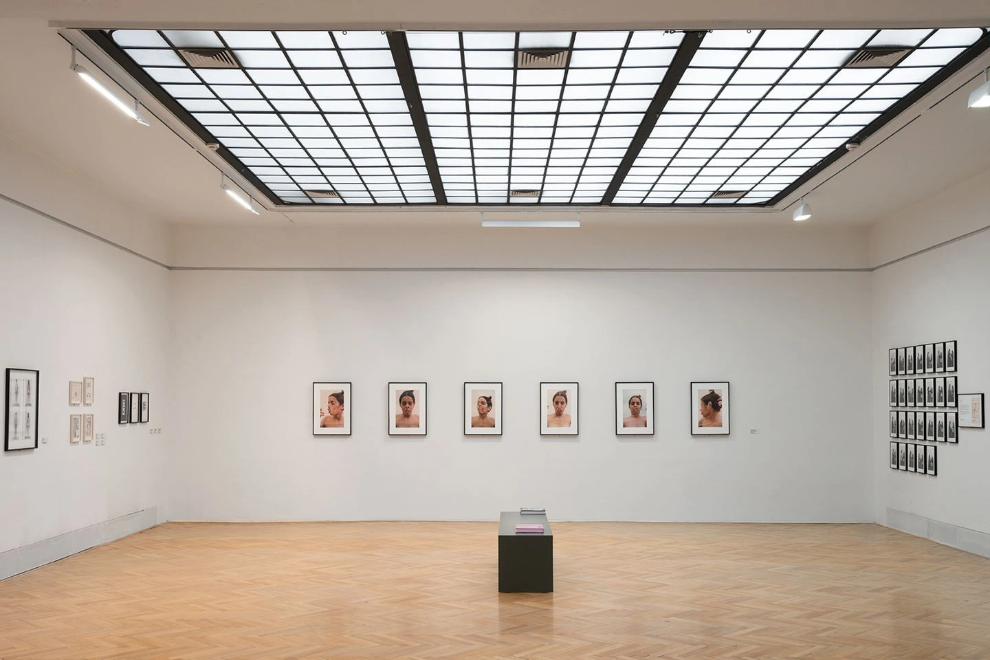 An exhibition room in the total view with wooden flooring can be seen. On the wall are portrait photos with black frames. Sunlight shines through the glass ceiling.