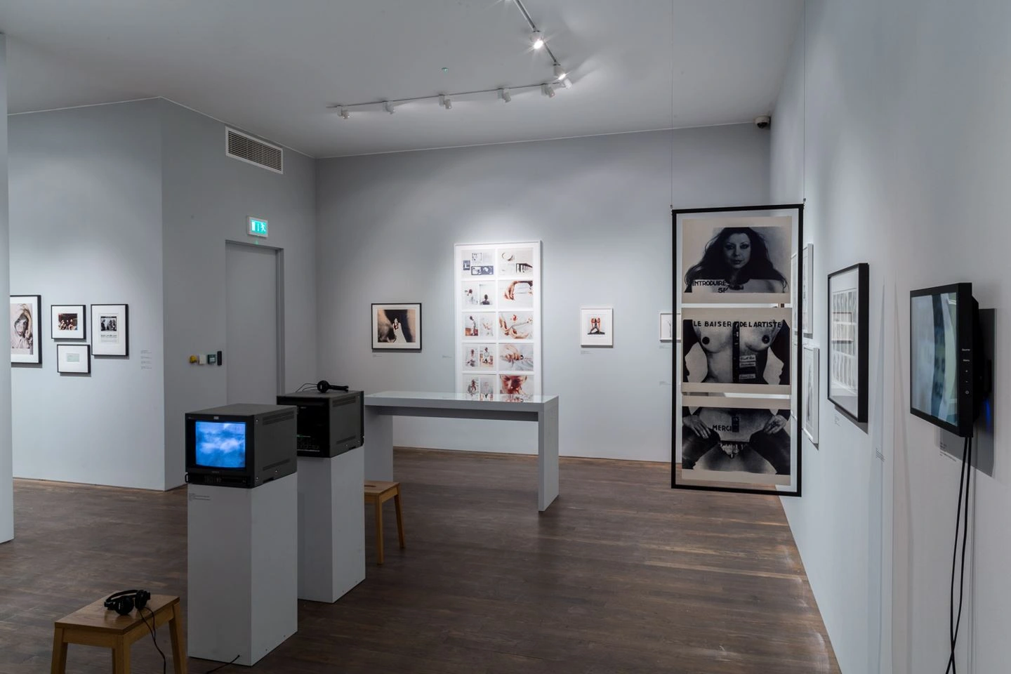 Interior view of an art gallery showcasing a variety of exhibits including framed black-and-white photographs on the walls, a vintage television installation on a plinth, and interactive multimedia displays on a dark wooden floor.