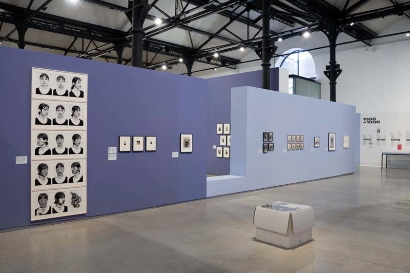 The picture shows part of the Feminist Avant-Garde exhibition in Arles. We see several exhibition areas presenting the different works. Many of them are black and white photographs. The room is divided by a black supporting structure. The walls are white.