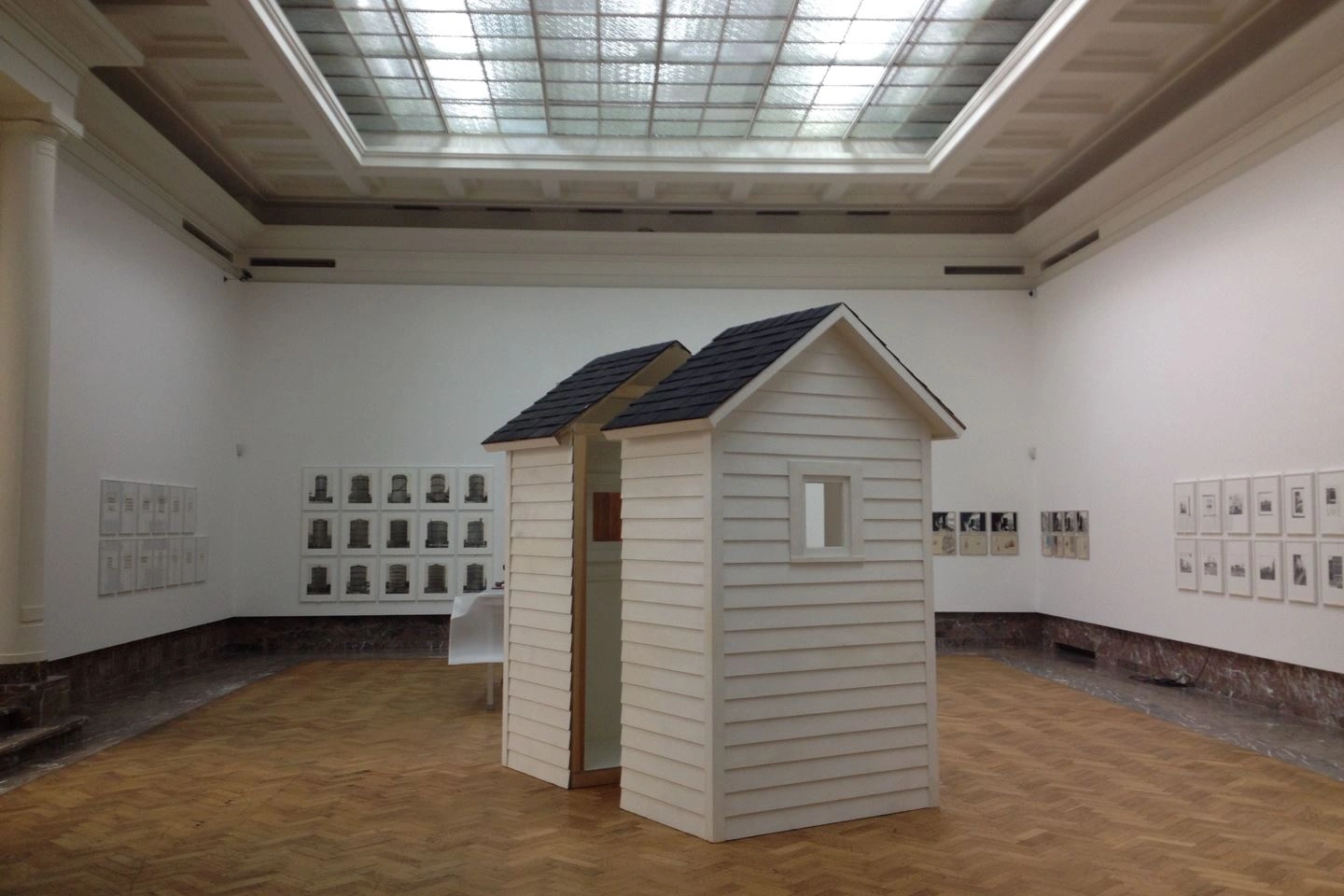 Installation view inside an art space featuring a small wooden house exhibit centrally placed, surrounded by paintings and graphic works on white walls beneath a skylight ceiling.