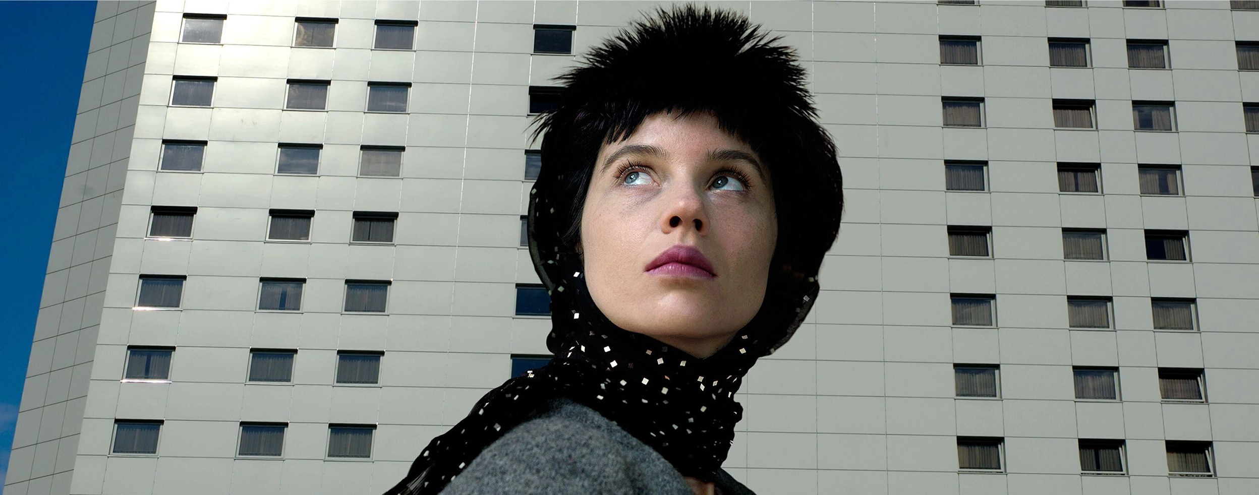 Aneta Grzeszykowska's photograph shows a woman with dark hair and a black scarf, which also partially covers her head. She has blue eyes and is looking upwards to the left. She is wearing gray and in the background you can see a simple skyscraper.