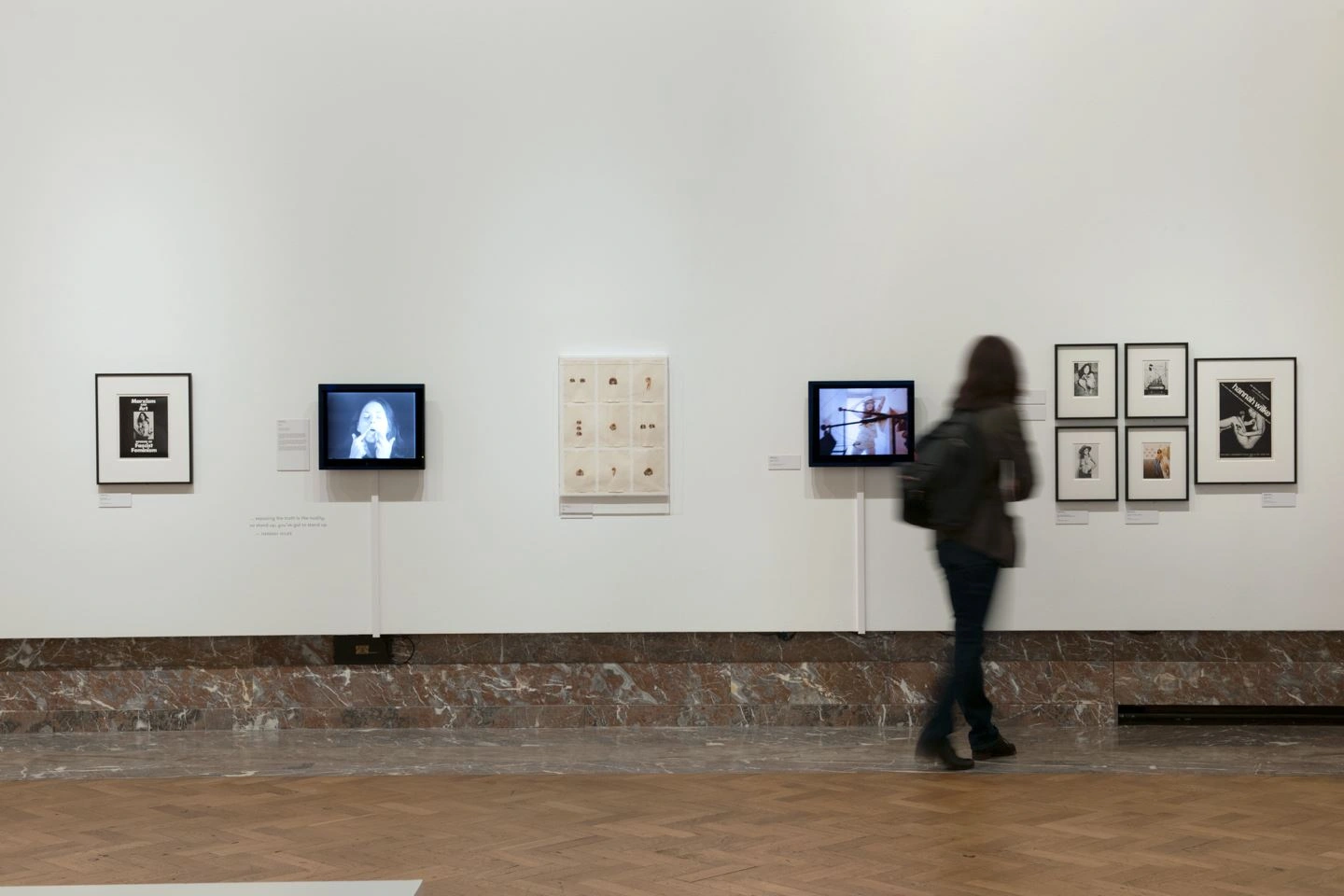 In einer Ausstellungshalle sind an einer weißen Wand mehrere Kunstwerke von Bozar aufgehängt. Ein Besucher betrachtet die Bilder und ist von hinten  zu sehen.