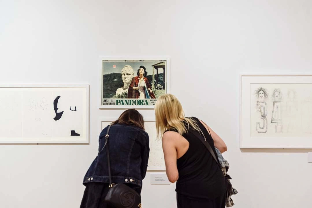 Two individuals closely observing a colorful movie poster labeled "PANDORA" amidst black and white artworks on a white gallery wall.