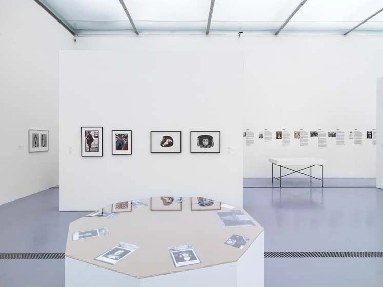 Ein sehr moderner und heller Ausstellungsraum zeigt an den Wänden Fotographien in schwarzen Rahmen. Im Raum stehen zwei Schautische. Einer davon spiegelt die Bilder an der Wand dahinter.