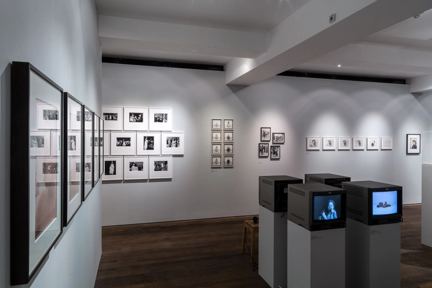 Innenansicht einer Fotokunstgalerie mit zahlreichen gerahmten Schwarz-Weiß-Fotografien an hellen Wänden und multimedialen Präsentationspodesten mit Bildschirmen in einer Ausstellungshalle mit Holzboden und moderner Beleuchtung.