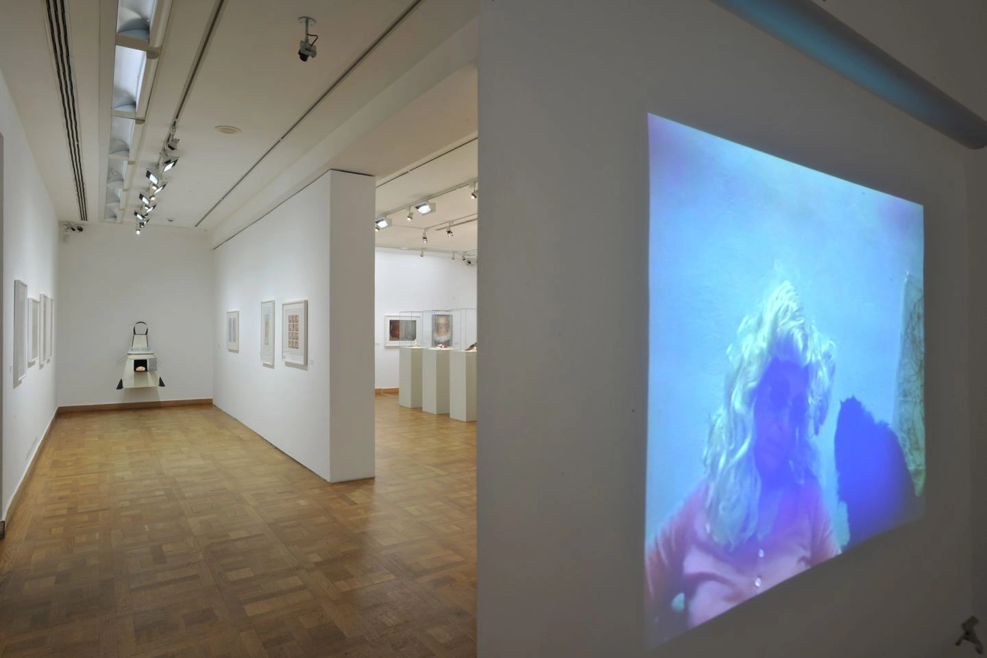 View inside a bright gallery with wooden paneled flooring, various framed pictures are hanging on the walls. At the end of the room, a large video artwork is projected onto a wall depicting a blurred figure underwater.