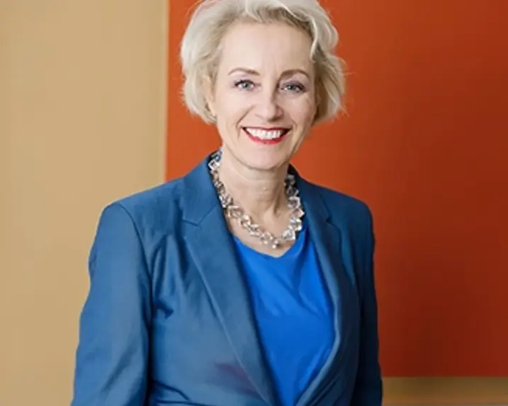 Gabriele Schor wears blue, smiles and stands in front of an orange background.