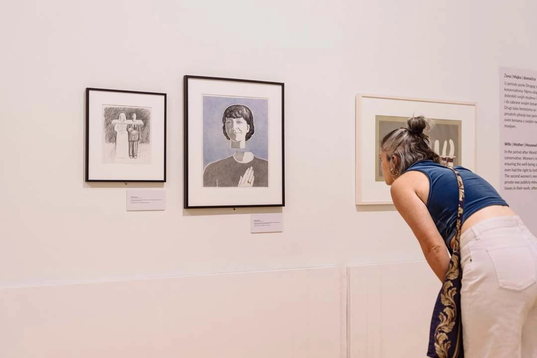 A gallery visitor leans forward to closely examine a detailed black-and-white portrait in a black frame, alongside other framed artworks on a light-colored wall.