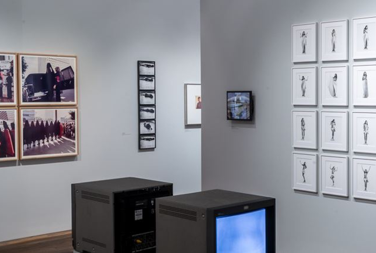 Interior view of an art gallery featuring a wall collage of framed black-and-white drawings, video installations on monitors, and abstract photographs in the background, prepared for viewing by gallery visitors.