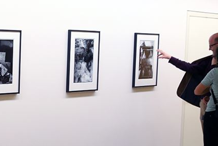 Zu sehen sind zwei Besucher der Ausstellung die vor  vier Fotographien auf einer weißen Wand stehen. Die Fotographien sind schwarz weiß und sind in einem passepartout Rahmen.