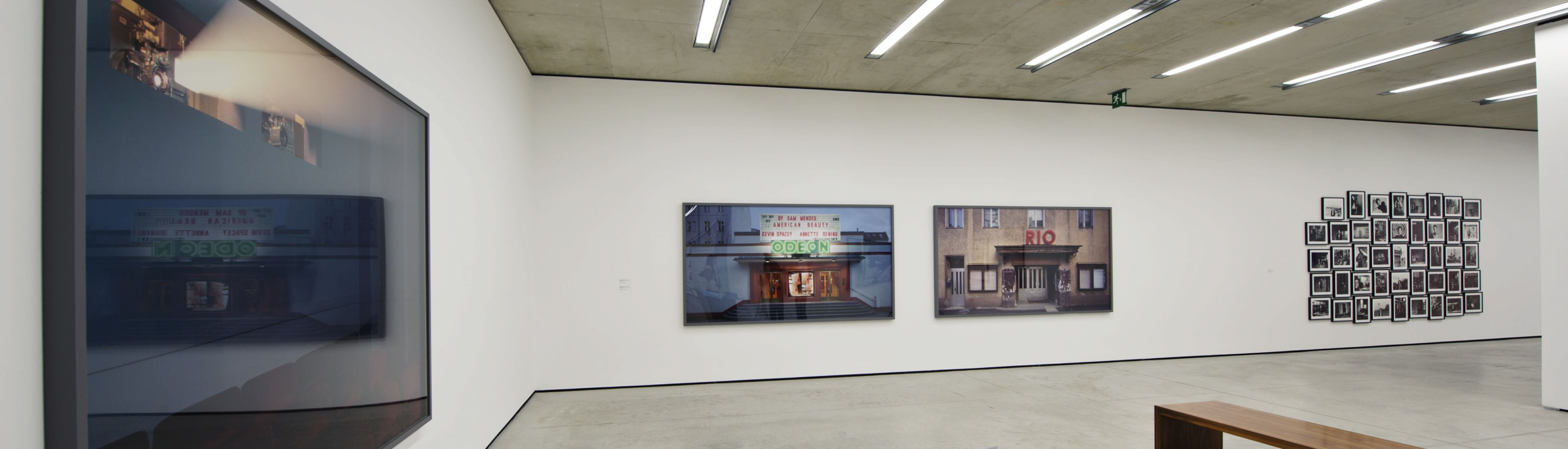 Bright gallery with two large cityscape paintings on a white wall and a wooden bench in the foreground.