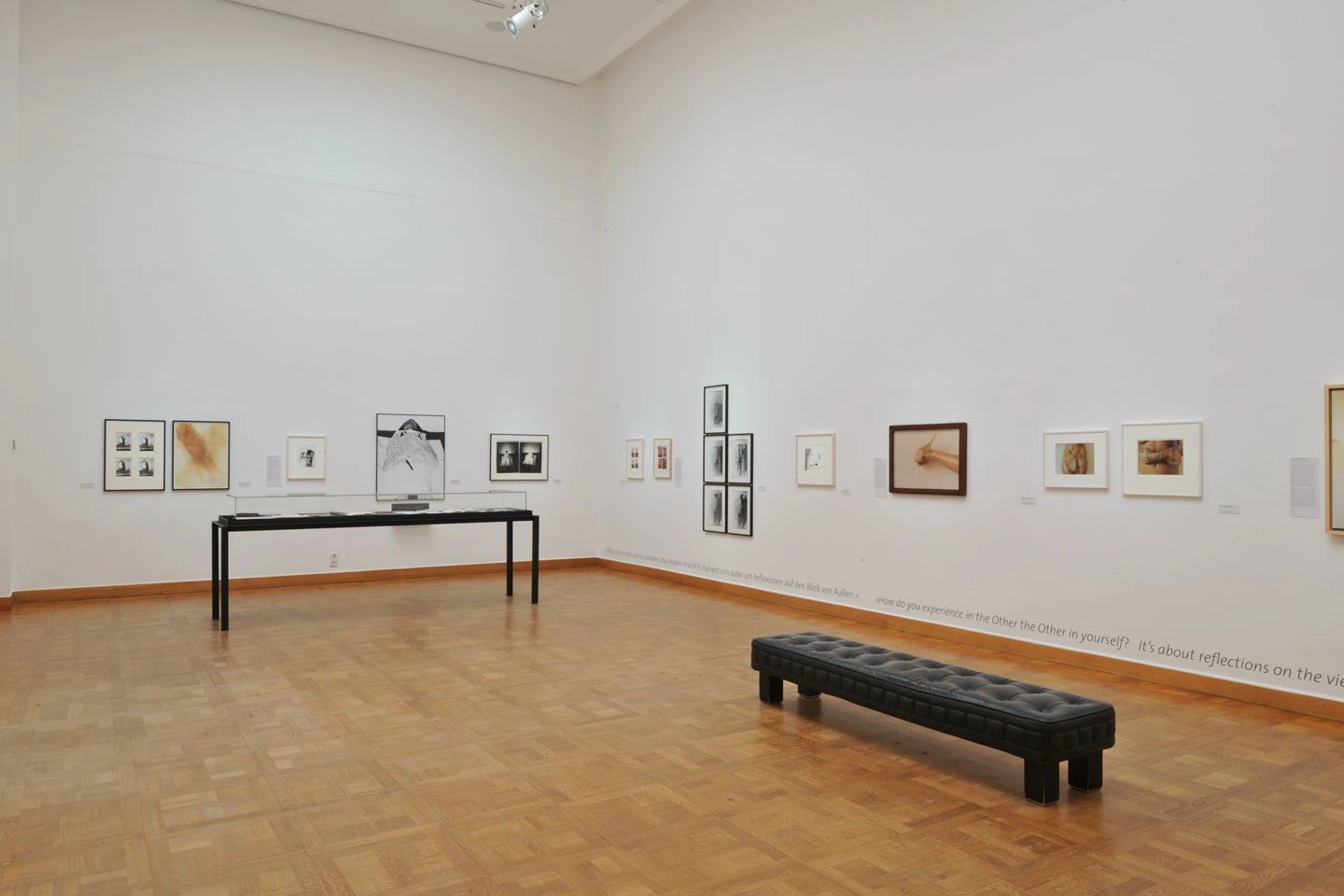 A modern art gallery with white-painted walls displays a collection of framed artworks of various sizes. A black upholstered bench is placed in the center of the room on a polished wooden floor.