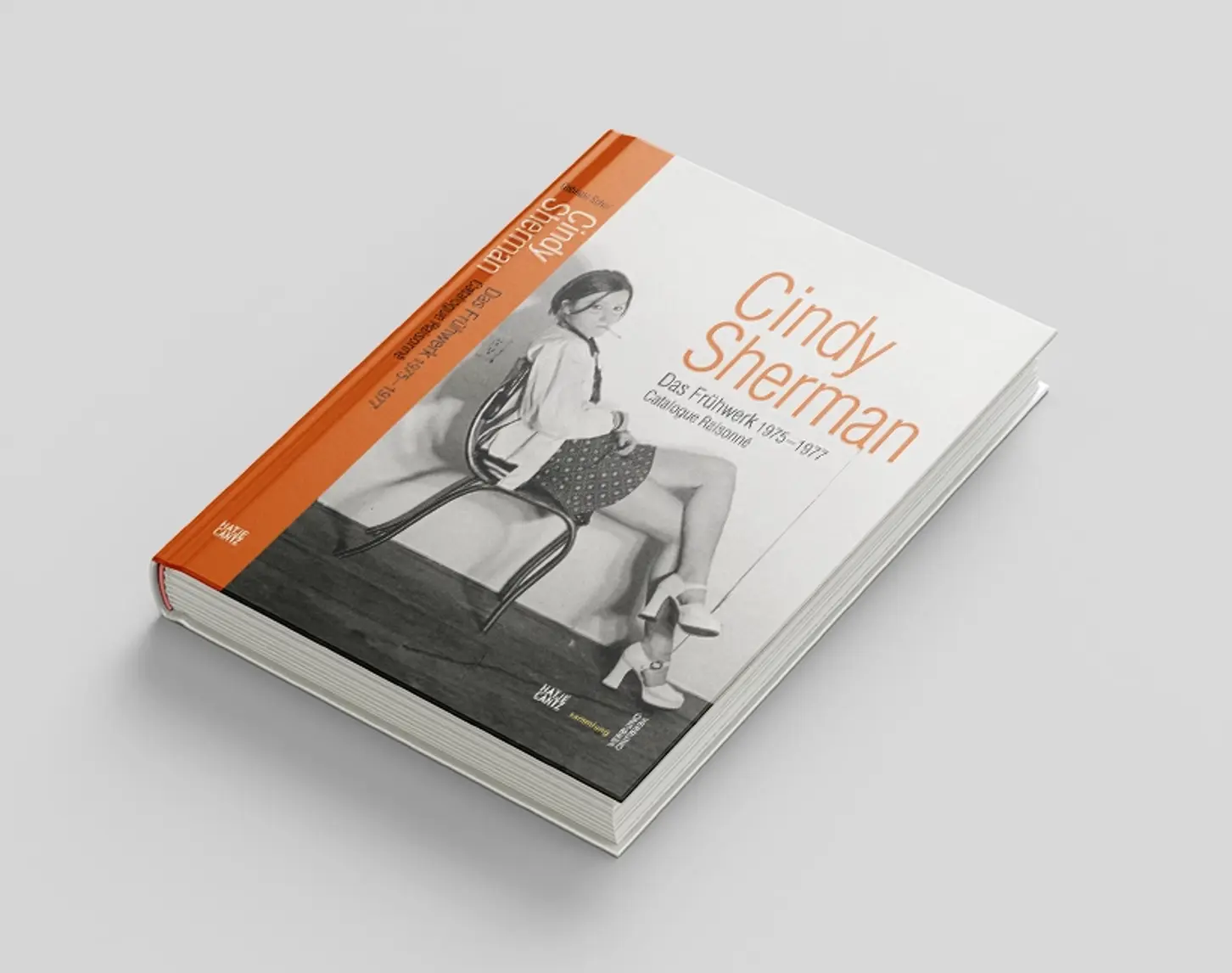 The exhibition catalog of Cindy Sherman's early work shows a photograph of a woman in black and white. She sits sideways on a chair, wearing a short skirt and a white top. She smokes. The font and the spine of the book are orange.