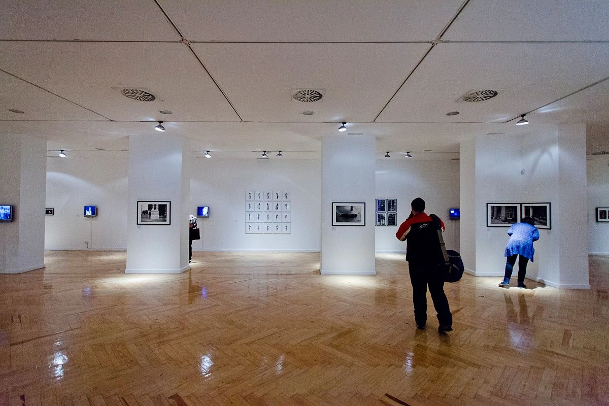 A close-up of a large exhibition room with white walls and a wooden floor can be seen. Several visitors are standing in the room looking at different pictures.