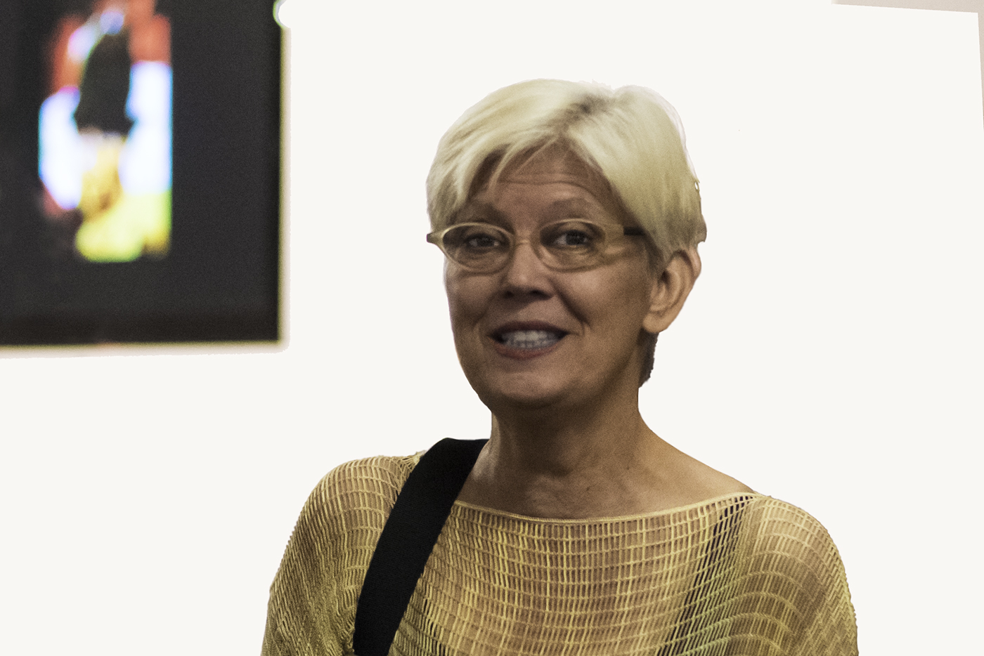 A portrait photo of the artist Loise Lawler can be seen. The artist wears short blond hair, narrow, light-colored glasses and a yellow top. Her bag hangs over her left shoulder.