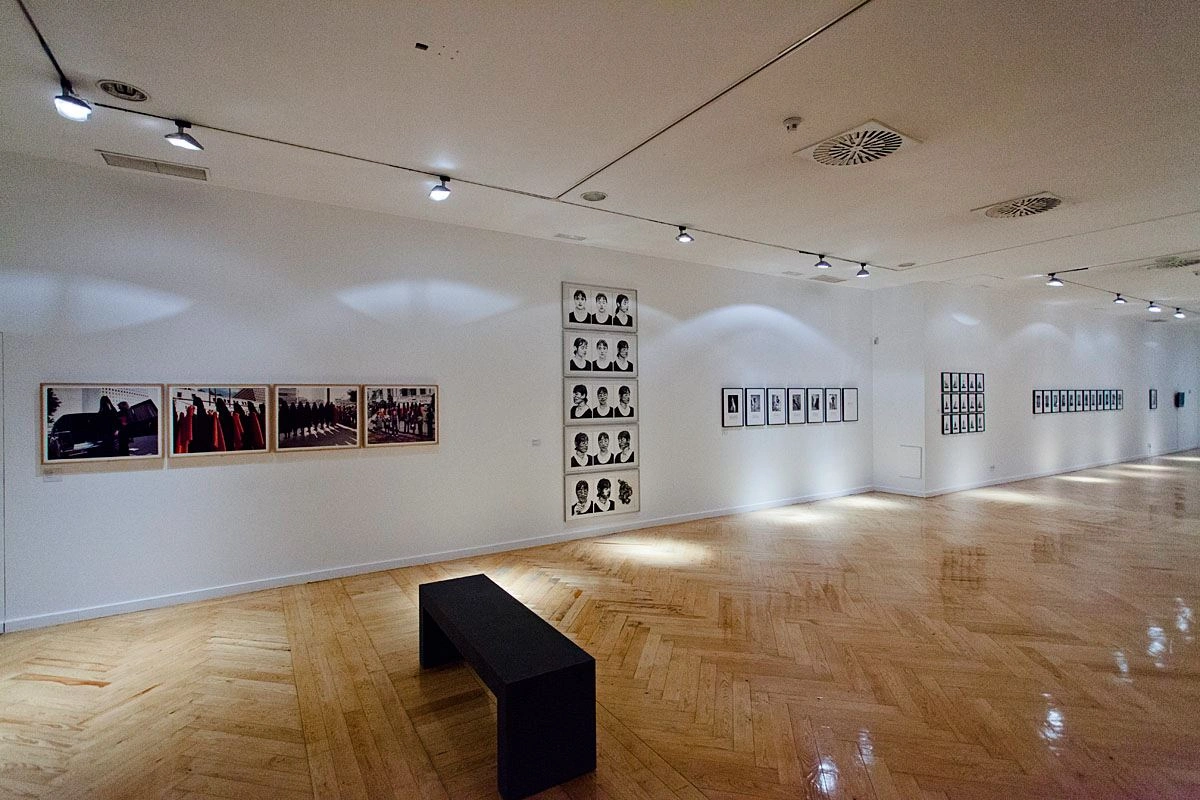 A modern exhibition room with white walls and a lacquered wooden floor. Several exhibits are hung on the wall and there is a black wall in the middle of the room.