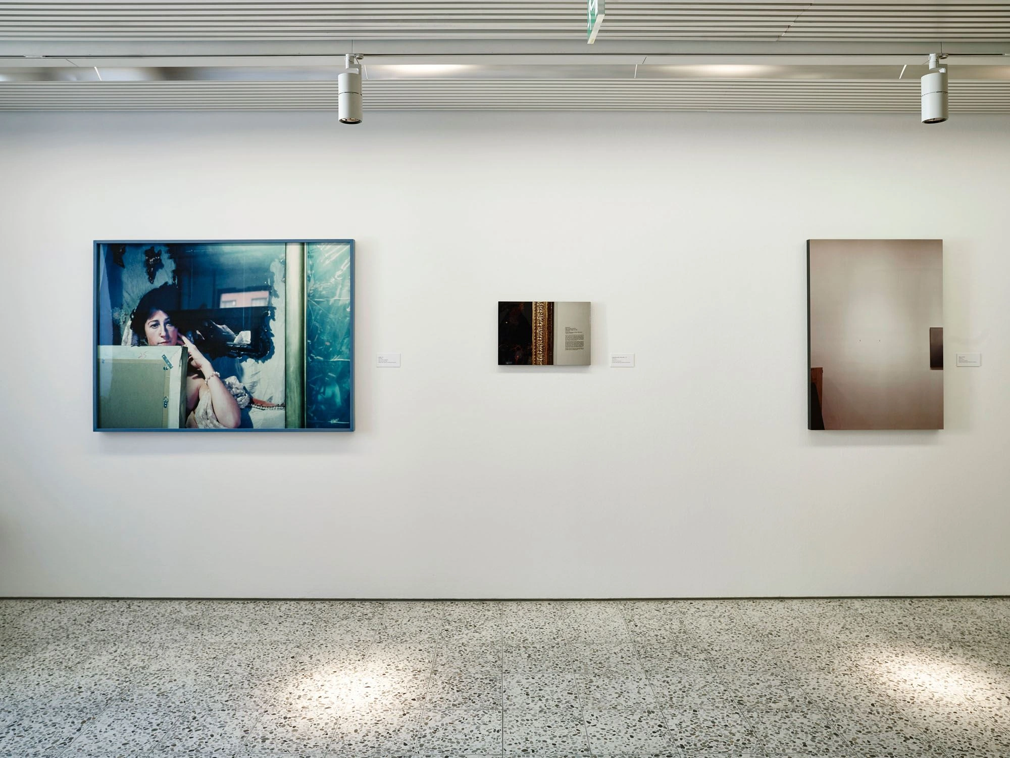 Three works of art hang on a white wall. To the left and right are large-format paintings and in the middle a small-format painting. Spotlights are placed on the ceiling that shine on the marbled floor.