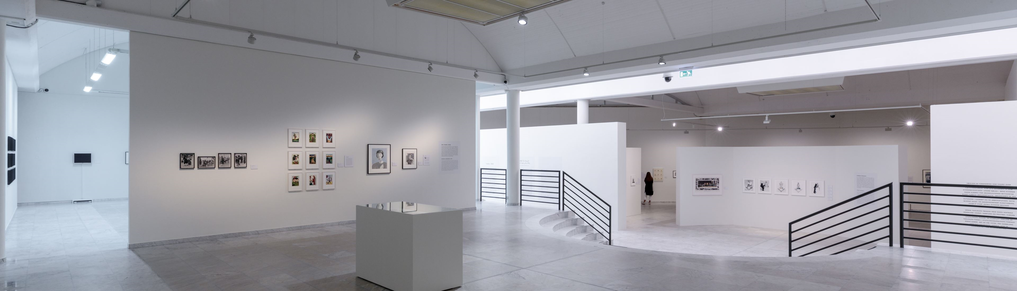 Eine Großaufnahme eines sehr hellen Ausstellungsraumes ist zu sehen. Rechts davon ist ein Treppengeländer. Der Ausstellungsraum ist mehrfach von Trennwänden geteilt. An den Trennwänden sind Kunstwerke aufgehängt. An der Decke des Raumes sind Lampen mit Spots befestigt.