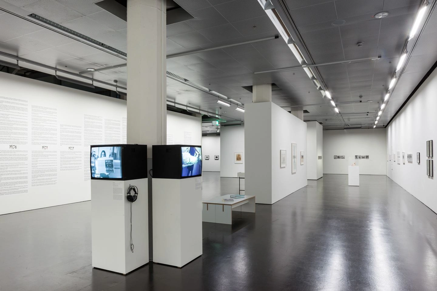 A large exhibition room divided by a partition wall can be seen. Works of art are depicted on all walls. In the front area of the room in the middle, two pedestals with monitors are set up that play different videos.