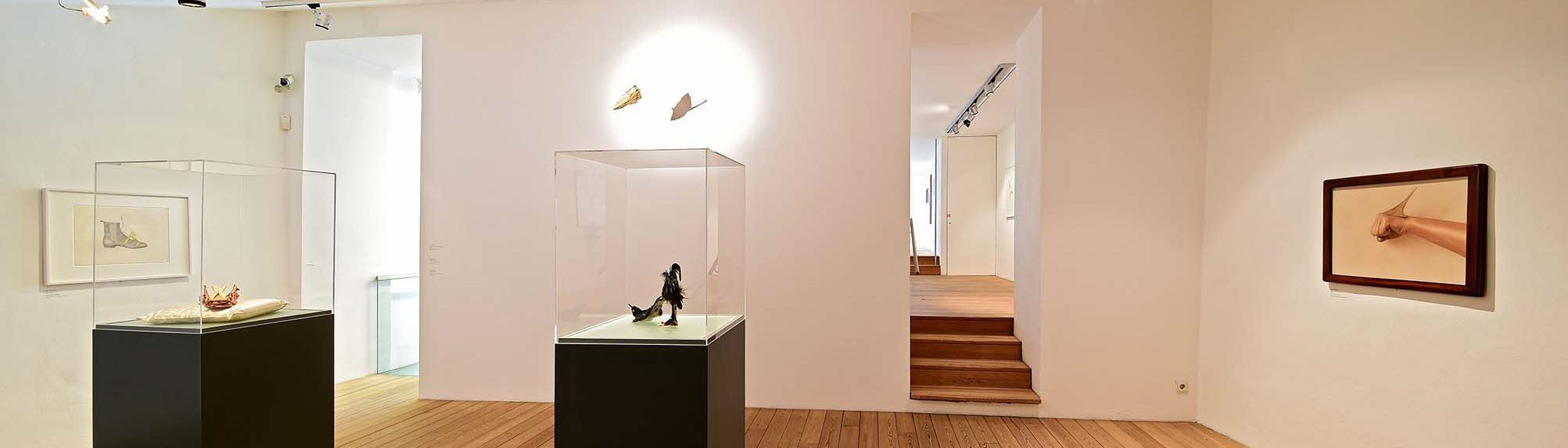 Panoramaansicht einer hellen Kunstgalerie mit Parkettboden, ausgestattet mit drei Vitrinen, die kunstvolle Objekte präsentieren, und einer gerahmten Wandzeichnung rechts, neben einer Treppe, die zu einem weiteren Stockwerk führt.