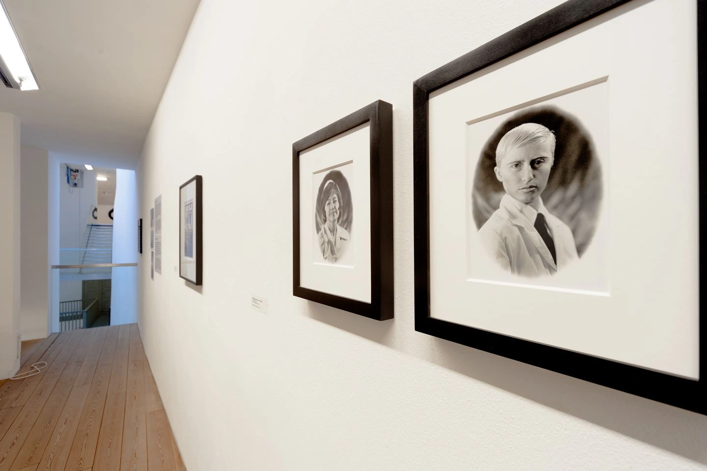 Two portrait photos hang on a white wall, on the right a young man with light hair and a white jacket, on the left a young lady with thinner hair.