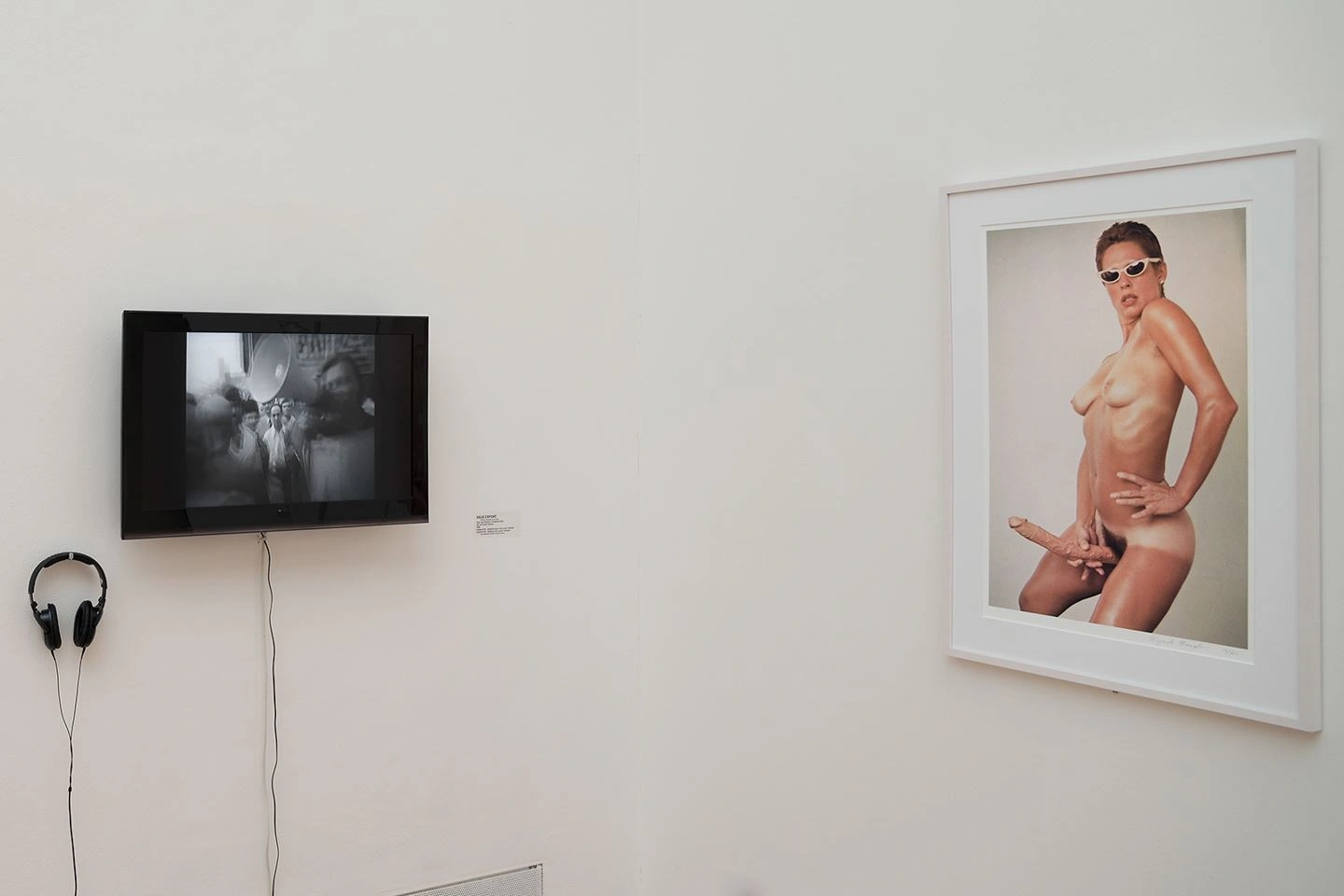 A monitor and headphones are mounted on one of the exhibition walls on the left. A video will play. On the right is a photograph in color on the wall in a white frame.