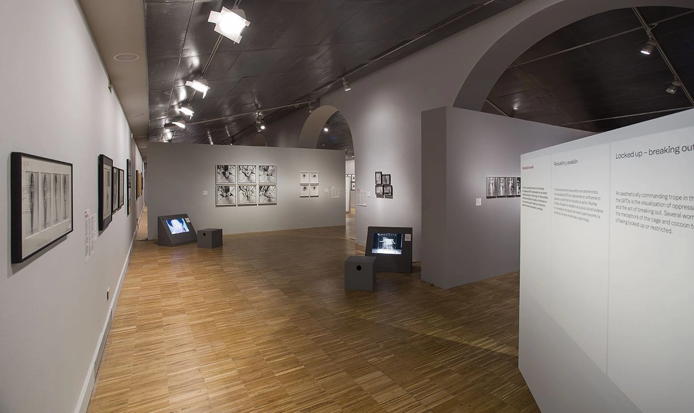 Interior view of a gallery with wooden floors and vaulted ceilings, walls adorned with framed artworks, multiple monitors on small stands displaying moving images, and informational text panels provide details about the exhibition.