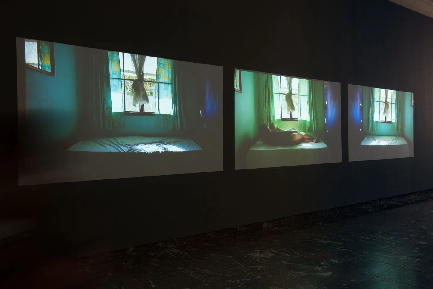 Three slide images are projected onto the wall on a black background in a dark room. Each shows a room with a mattress in front of a window in the curtain. In the middle image, two people are lying on the mattress.