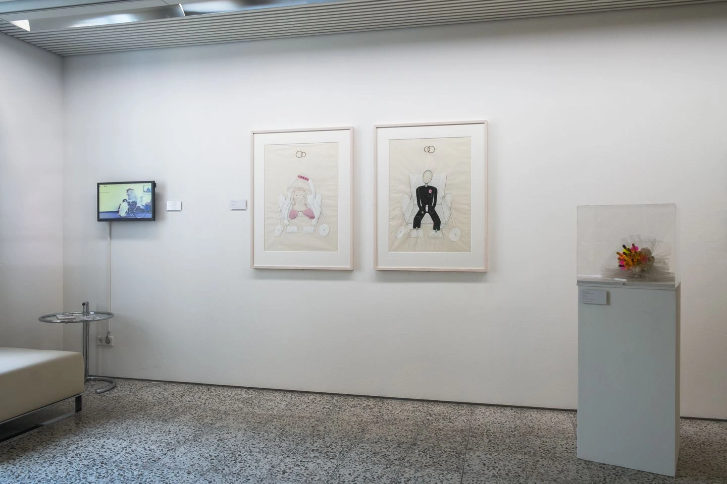 Bright exhibition space with two framed drawings on the wall, a multimedia installation on the left side, and a display case with flowers on the right.