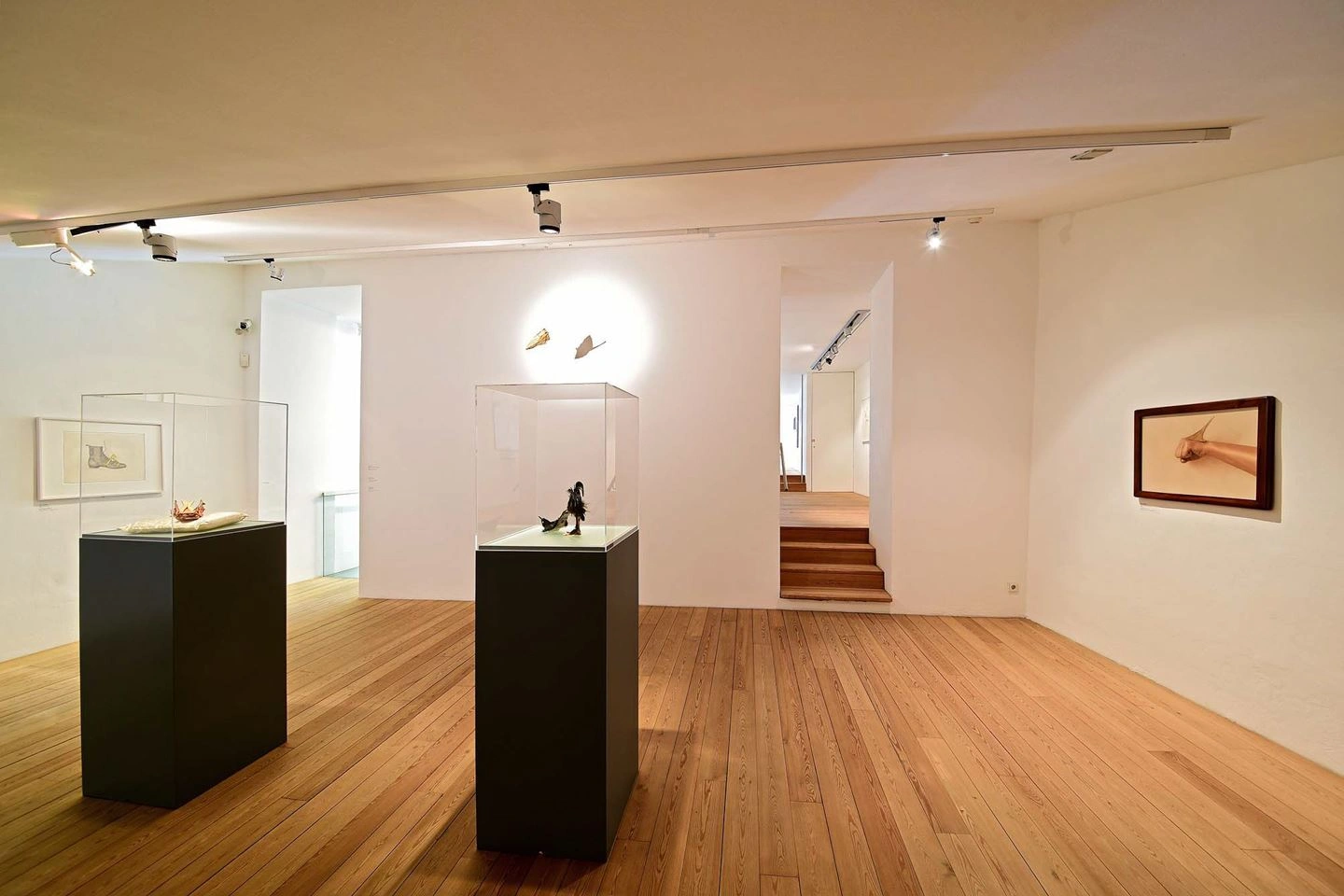 Interior view of an art gallery with wooden parquet flooring and white walls, displaying various works of art. Two sculptures are set on black pedestals in the center of the room, with stairs leading to another level in the background.