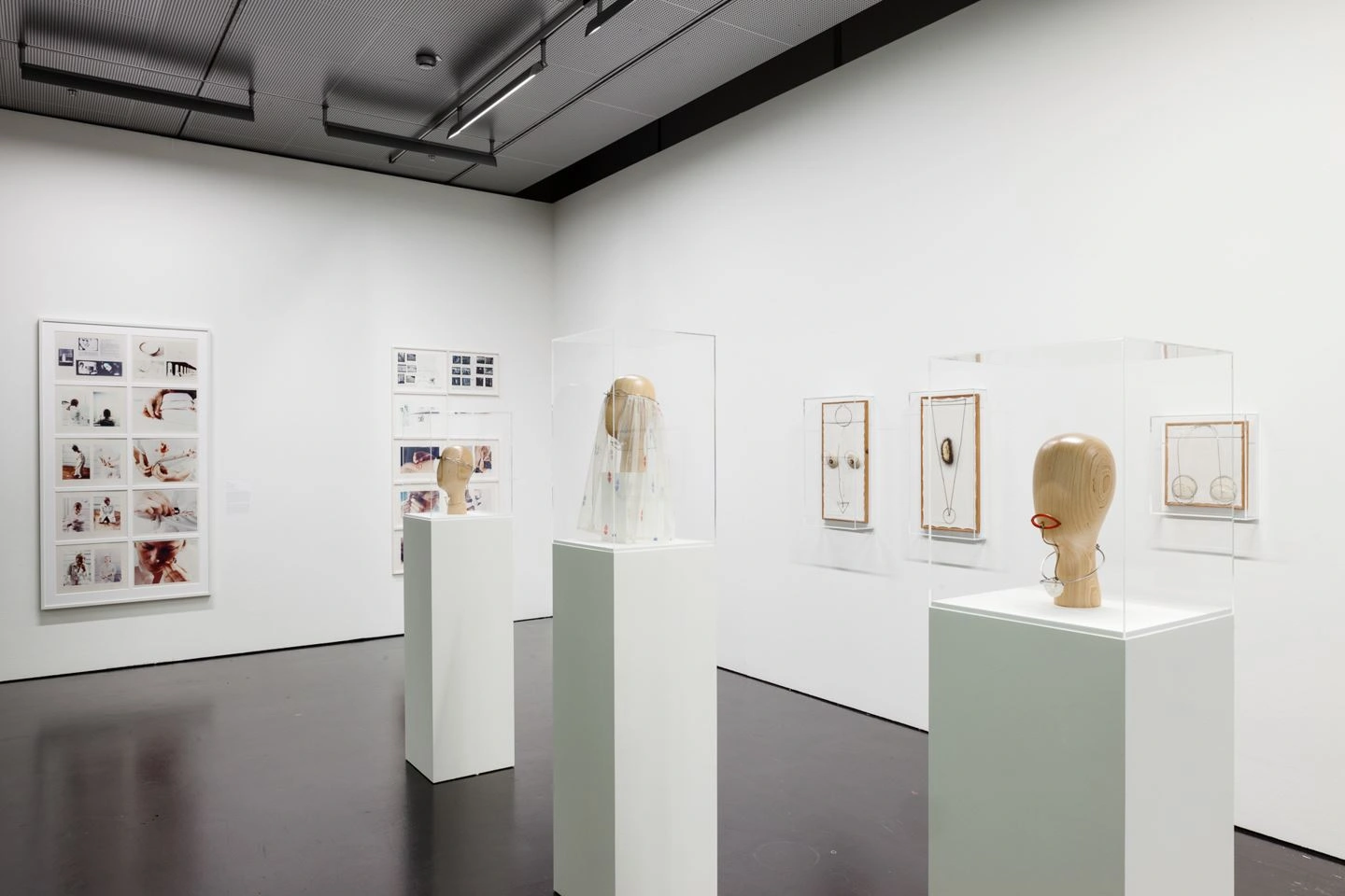 A modern exhibition room with white walls and dark floor can be seen. Several works of art hang on the walls and there are three showcases in the middle of the room