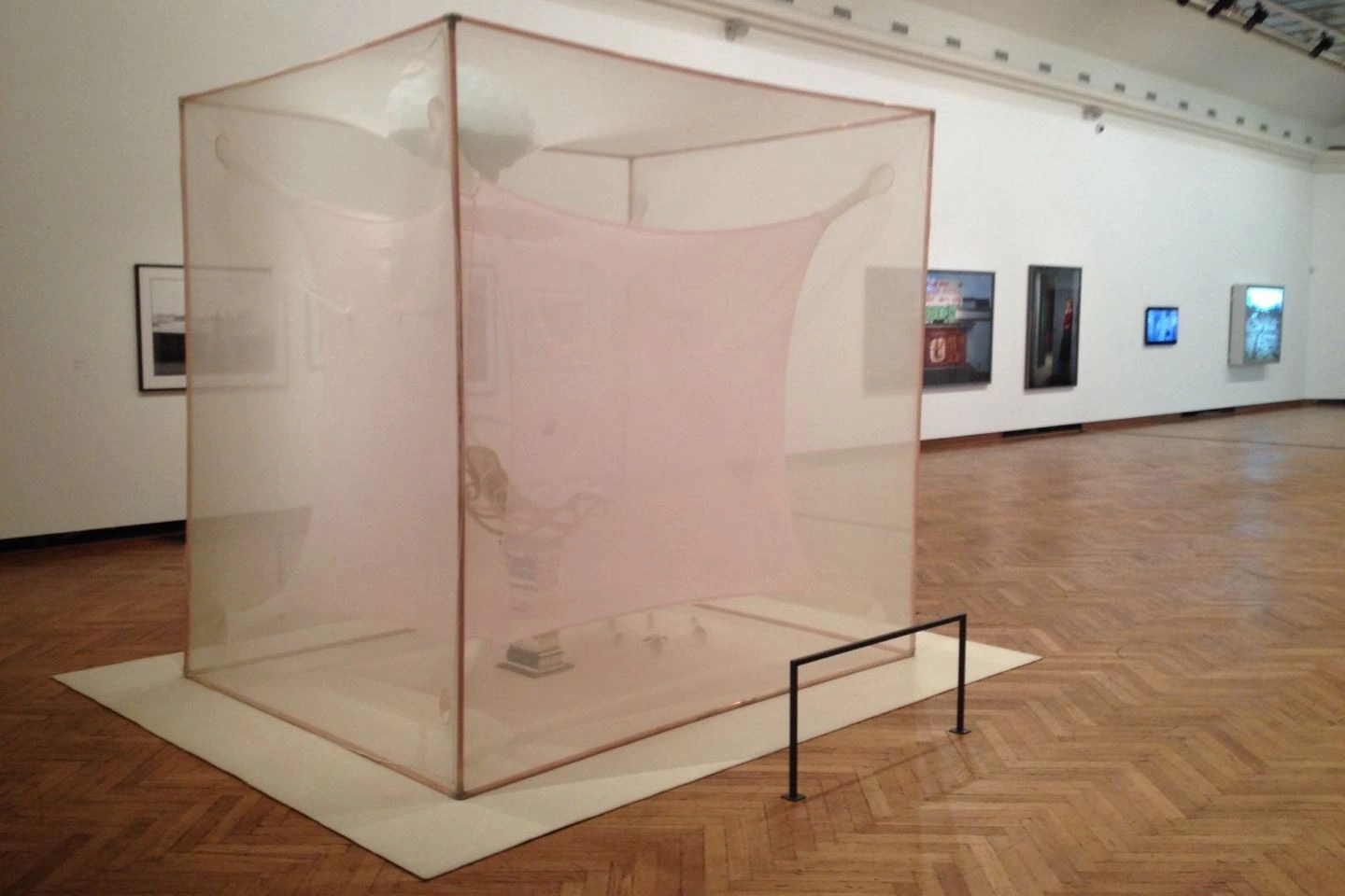 Transparent plastic box-shaped sculpture in the center of an art gallery, with paintings and photographs on the walls in the background, on parquet flooring.