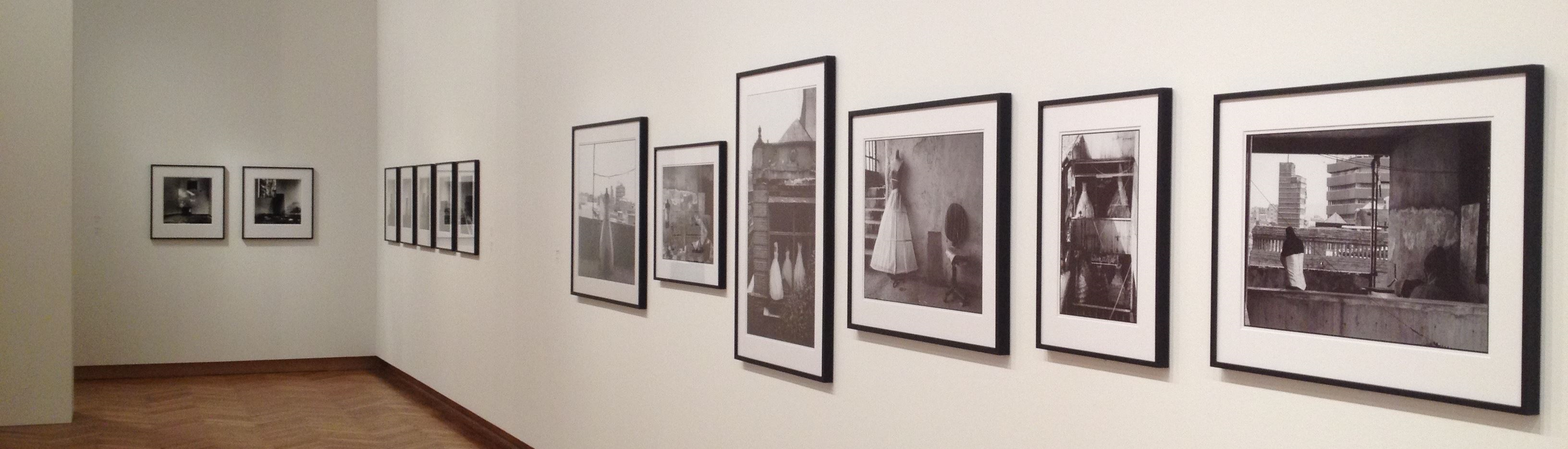 Interior view of a photo gallery featuring a selection of framed black and white photographs hung in chronological order on a white wall, in a room with wooden flooring and soft lighting.