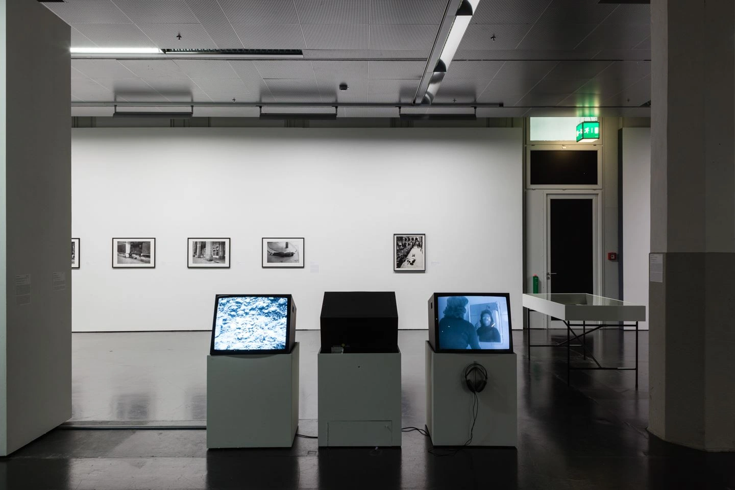 An exhibition room with dimmed light can be seen. Three monitors are set up in the middle. Two of them play a video. The monitor in the middle is turned off.