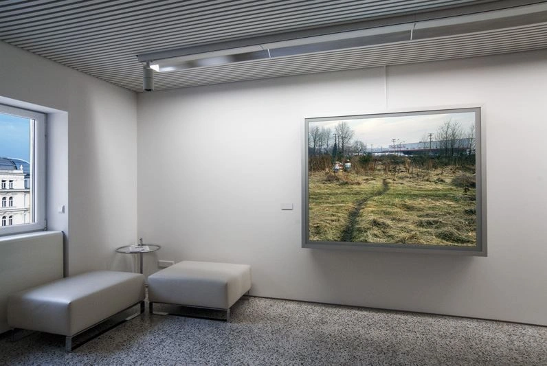 A modern office corridor can be seen with a corner bench and a corner window. Next to it is a photograph in a silver frame of a green meadow landscape.