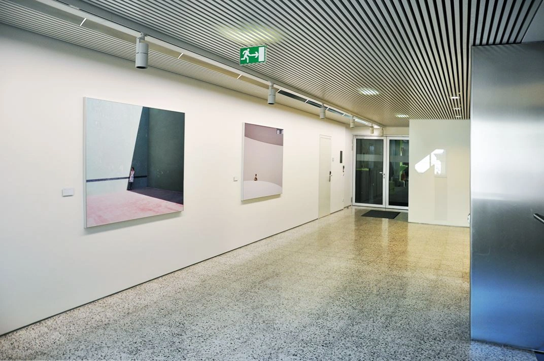 Bright, modern gallery hallway featuring three large-scale paintings on a white wall, terrazzo floor, and the entrance to another room visible in the background.