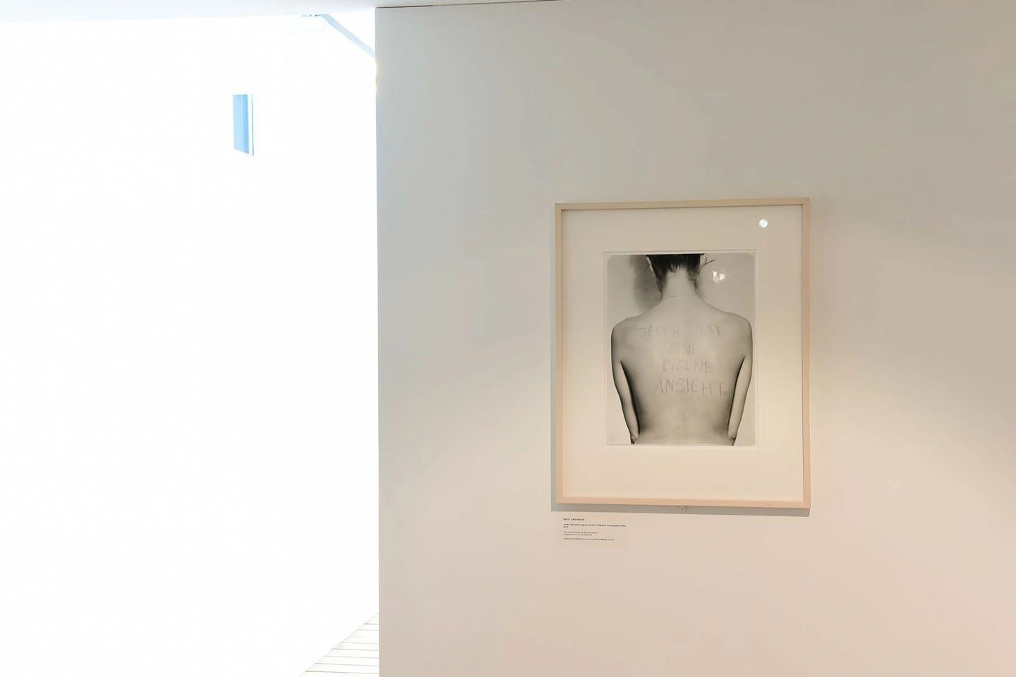 Black-and-white photograph of a human back, framed and illuminated on a gallery wall with an informational label underneath, in a brightly lit room with a white wall.