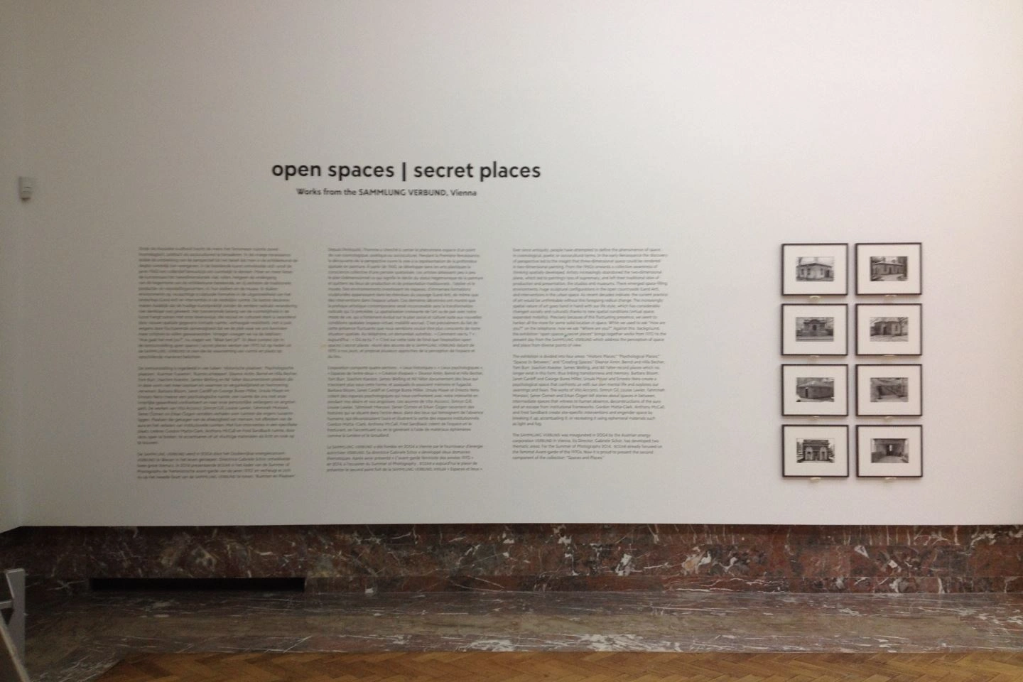 Exhibition view titled "open spaces | secret places" featuring text panels and a series of framed black-and-white photographs on a white gallery wall with a visibly rustic floor.