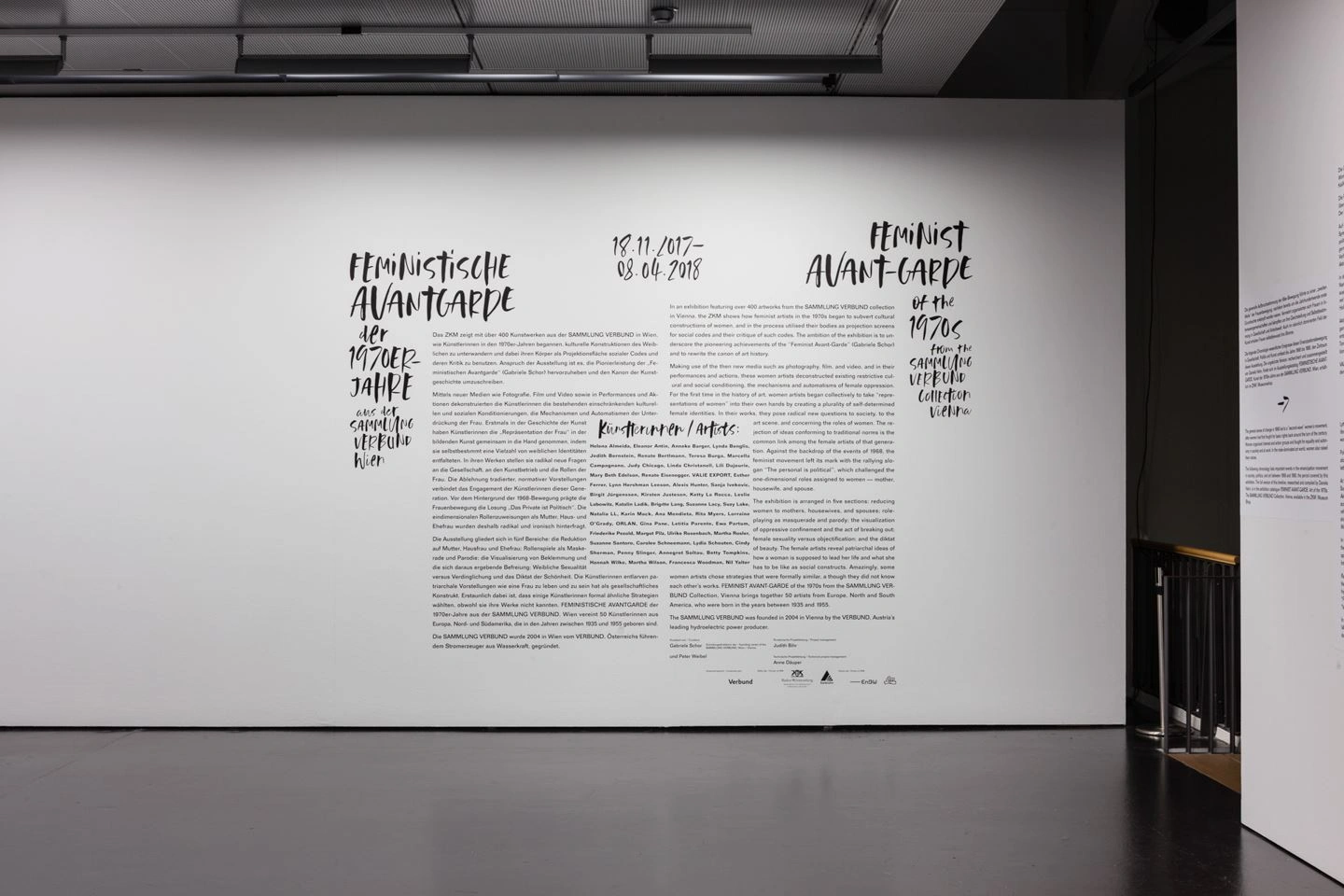 Ein Ausstellungsraum mit einer weißen Wand und dunklem Boden ist zu sehen. Auf der weißen Wand ist mit schwarzer Schrift ein langer Text zu sehen zum Thema Feministische Avantgarde.