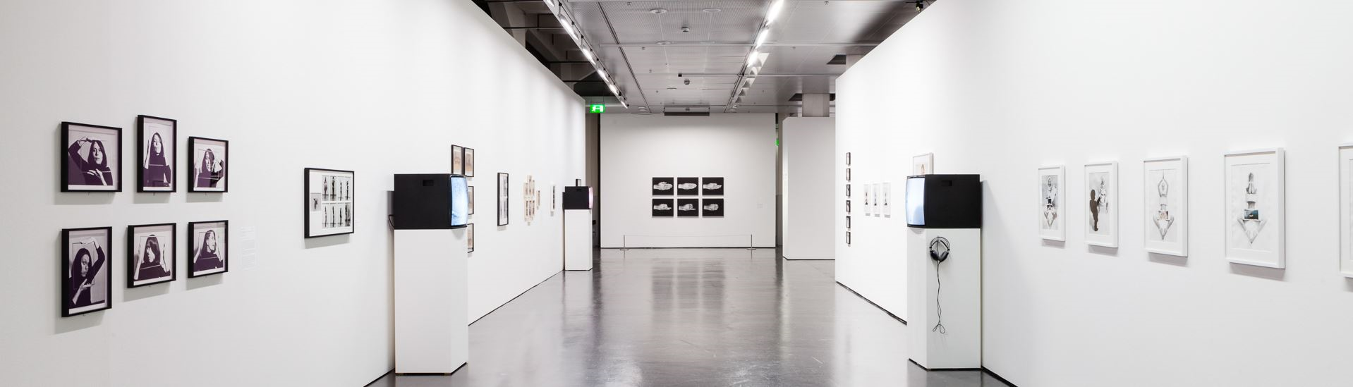 Ein langer Flur ist zu sehen mit Bildern auf weißen Wänden links und rechts. Links sind Portraitfotos in schwarz weiß mit schwarzem Rahmen und rechts Fotos von Personen in weißem Rahmen. Im Raum stehen außerdem Bildschirme, die Videos abspielen auf einem weißen Sockel.