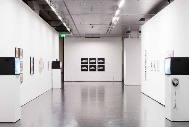 A long corridor can be seen with pictures on white walls left and right. On the left are portrait photos in black and white with black frames and on the right are photos of people in white frames. There are also screens in the room that play videos on a white base.