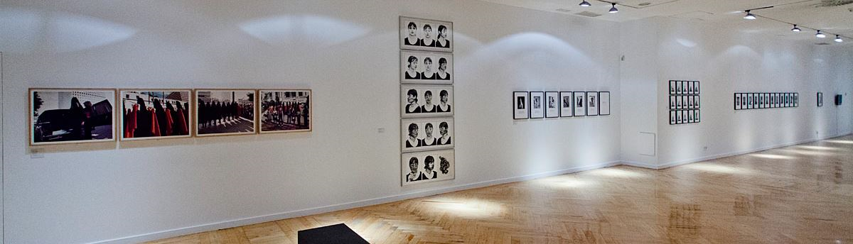 Ein moderner Ausstellungsraum im Panoramawinkel ist zu sehen. Auf der breiten Wand sind zahlreiche Kunstwerke zu sehen. An der Decke sind Lichtspots, die auf die Kunstwerke leuchten.