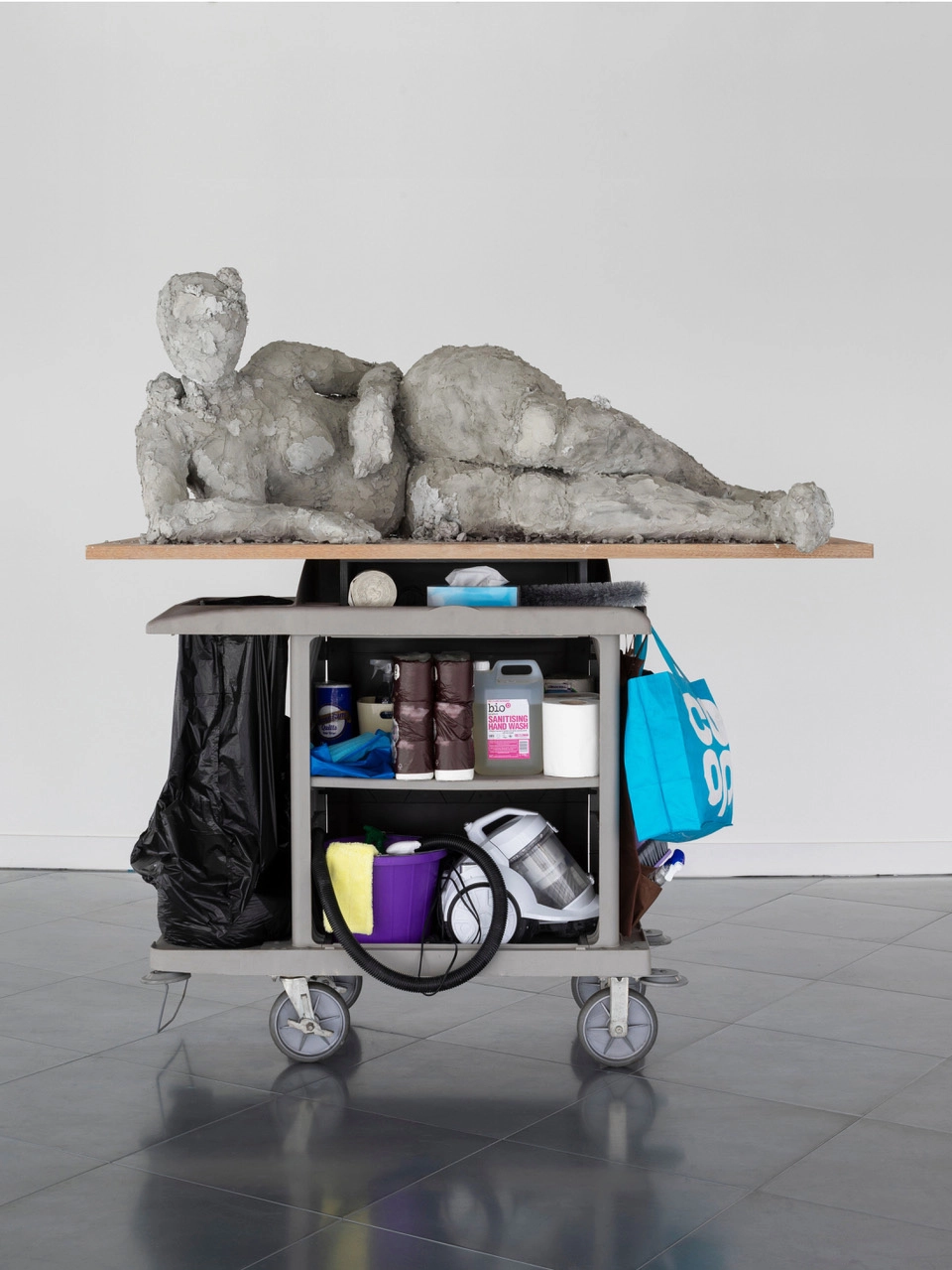 Wir blicken auf ein Kunstwerk von Nicole Wermers. Auf einem Reinigungswagen ist ein Holzbrett angebracht auf dem sich eine steinerne weibliche Figur zu sehen.