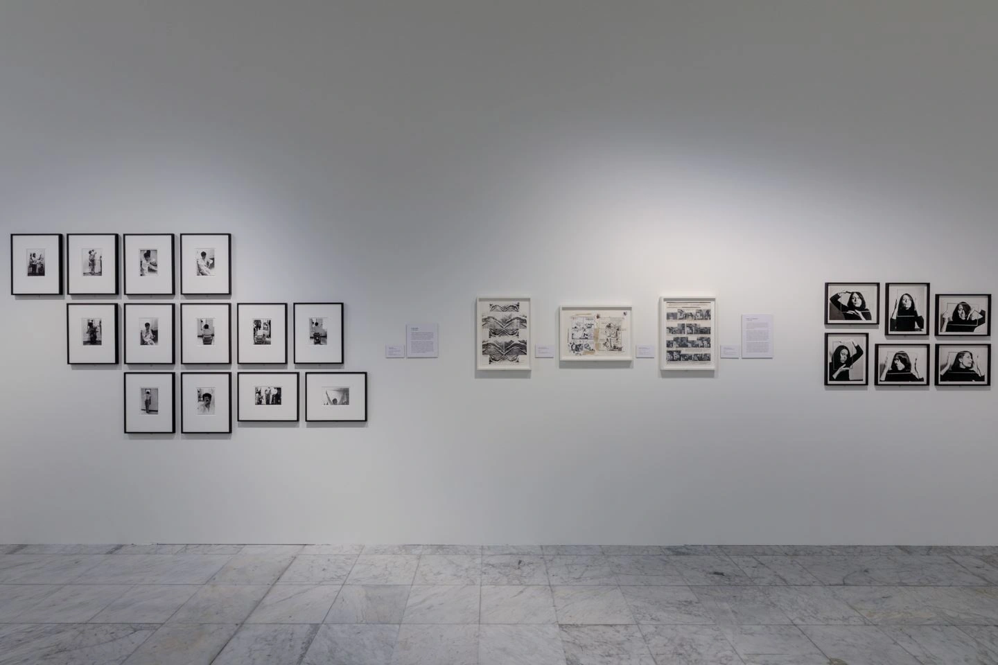 a somewhat darkened exhibition room can be seen. Several black and white photographs are hung on a white wall. On the far right are 6 portraits visible.