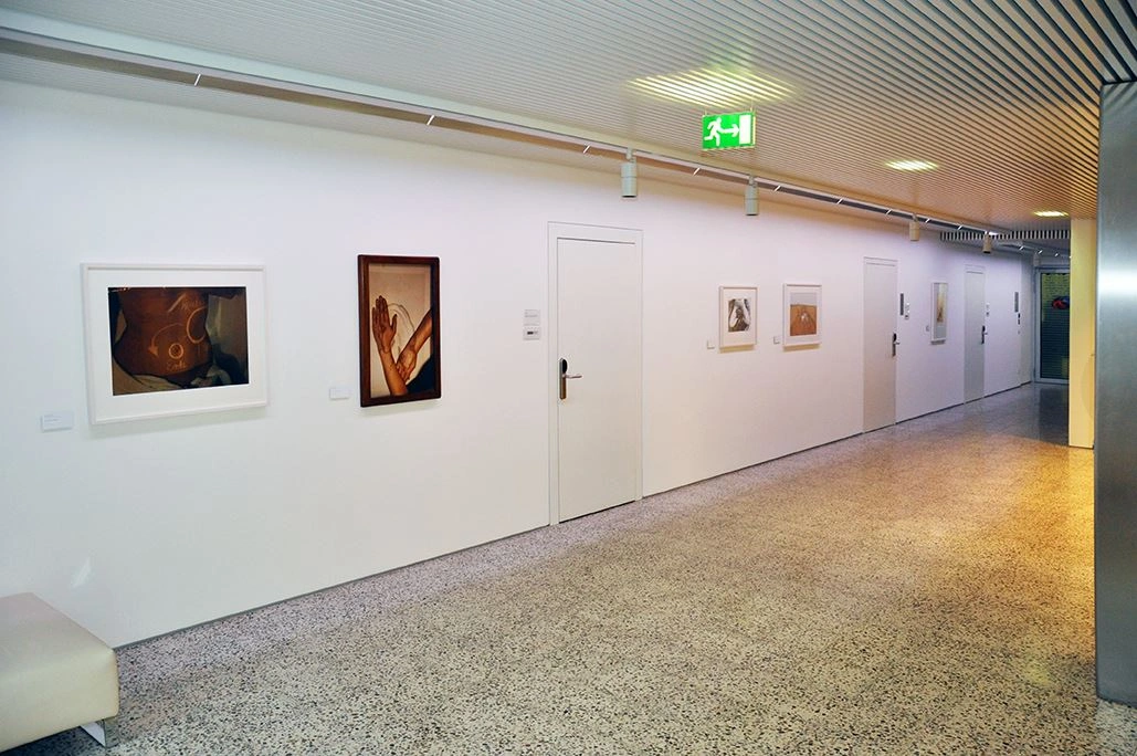 Hallway of an art gallery displaying framed paintings on a white wall, terrazzo flooring, and a bench on the left edge provides a space for resting.