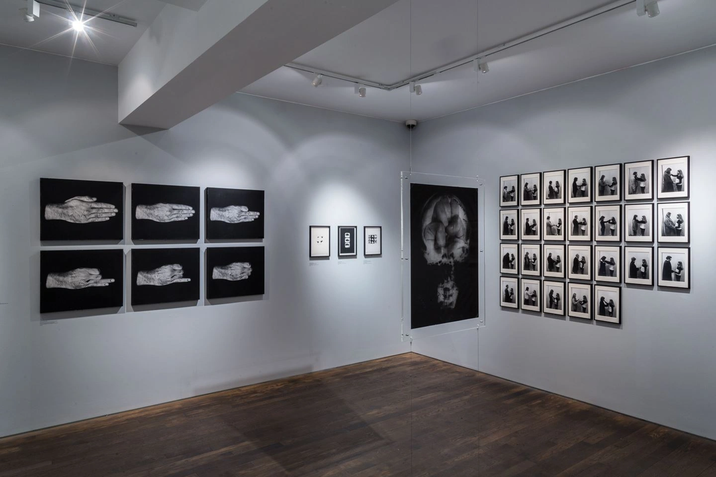 Corner of an art gallery exhibiting black and white artworks in various frames, including a series of abstract shapes on the left and portrait photographs on the right, on wooden flooring under ceiling spotlights.