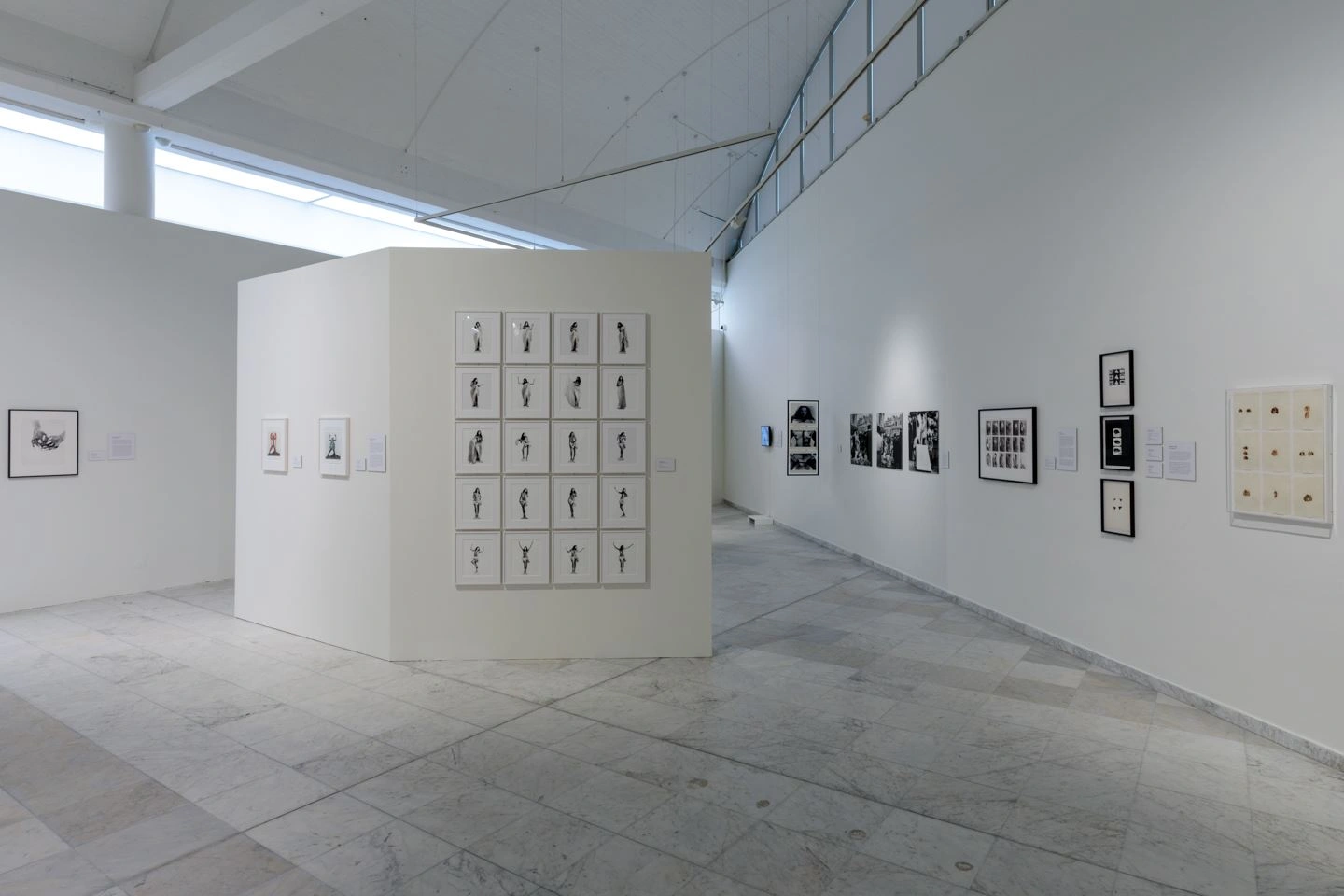 A high exhibition room with a light floor and white walls can be seen. On a partition wall is a series of black and white photographs. 5 rows of 4 photos each in a white frame.