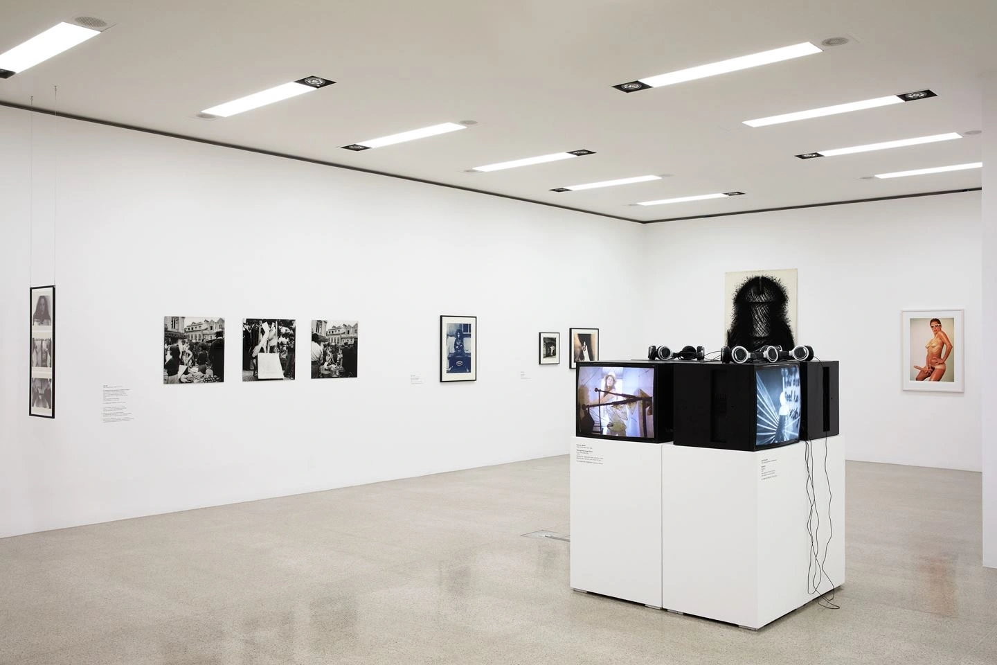 Zu sehen ist eine Totalaufnahme eines sehr hellen und kalt beleuchteten Austellungsraum. An der Wand sind unterschiedliche Fotographien aufgehängt und in der Mitte des Raumes befindet sich eine Kunstinstallation bestehend aus mehreren Monitoren, die ein Video abspielen.