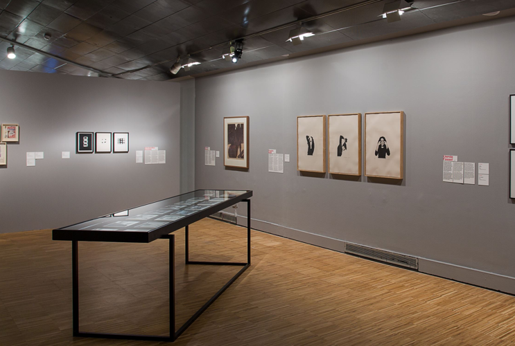 Cozy art gallery with light wooden flooring and gray walls, featuring a selection of framed drawings and photographs of varying sizes, lit by ceiling spotlights, and a glass showcase in the foreground presenting additional artworks and information.