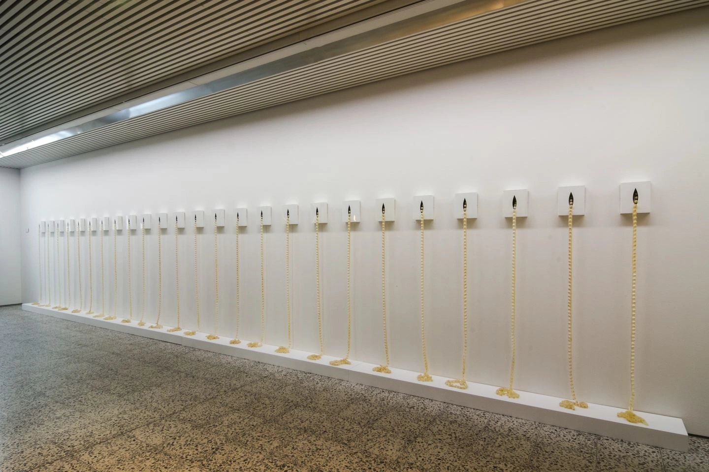 Art installation featuring a series of small, illuminated exhibits along a lengthy white wall in a gallery with textured lighting and terrazzo flooring.